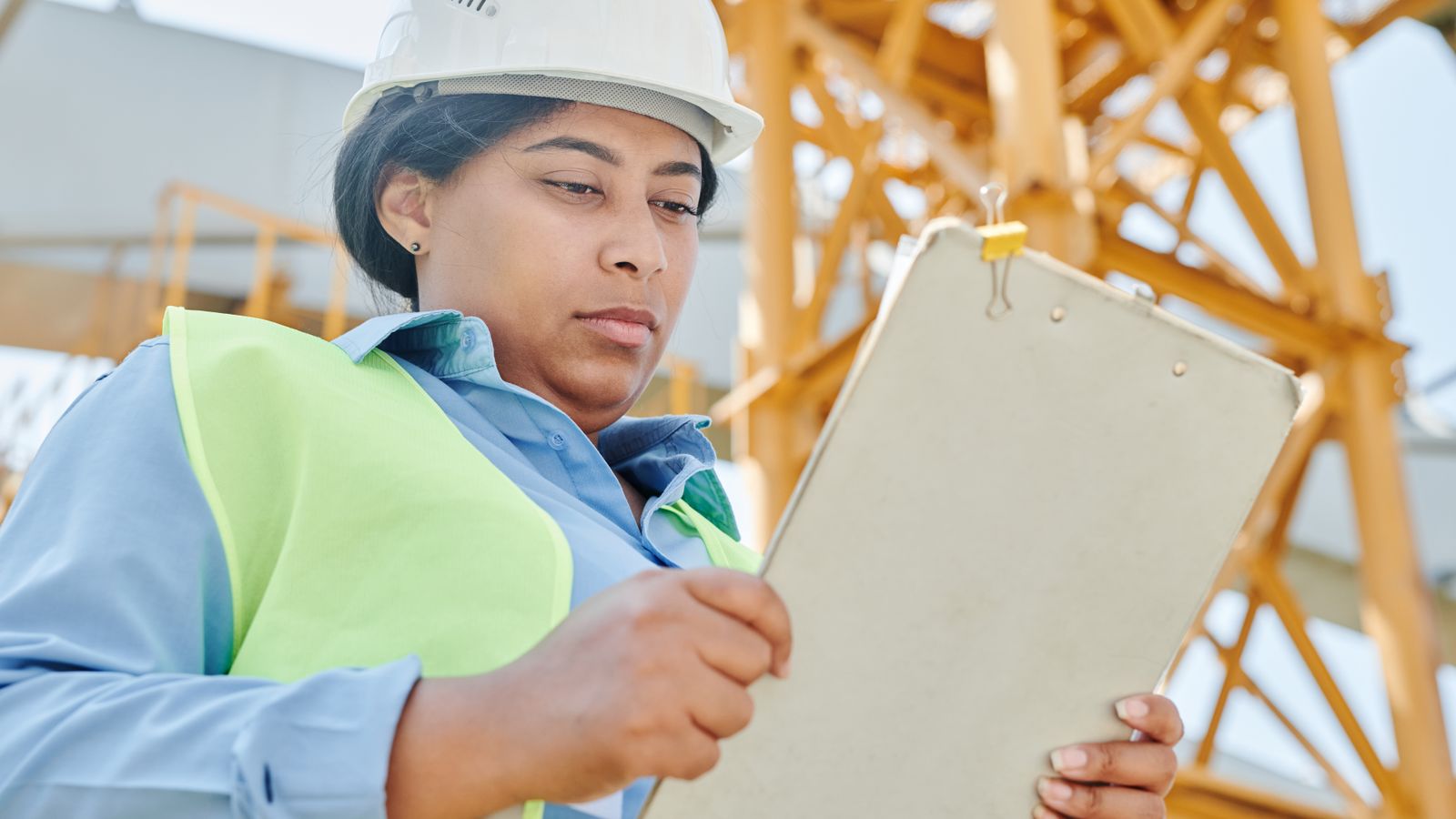 A woman in a hard hat and high-visibility vest studies a clipboard at a construction site