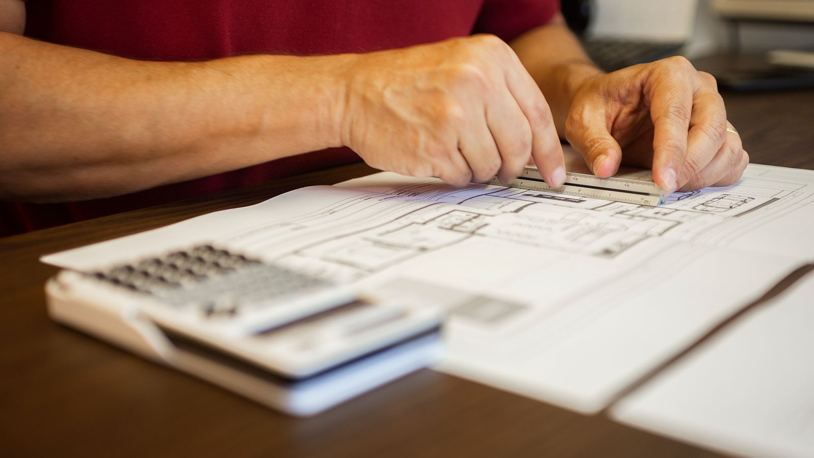 Hands measuring a floor plan with a ruler beside a calculator on a wooden desk