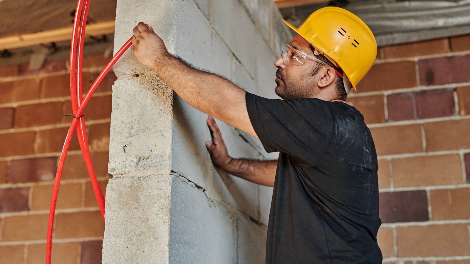 Un ouvrier casqué, avec lunettes de sécurité, installe un tuyau rouge sur un mur de blocs