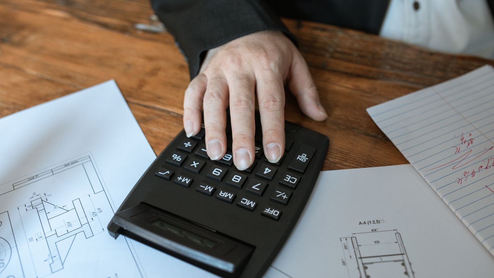 A hand rests on a calculator surrounded by technical drawings and an annotated sheet.