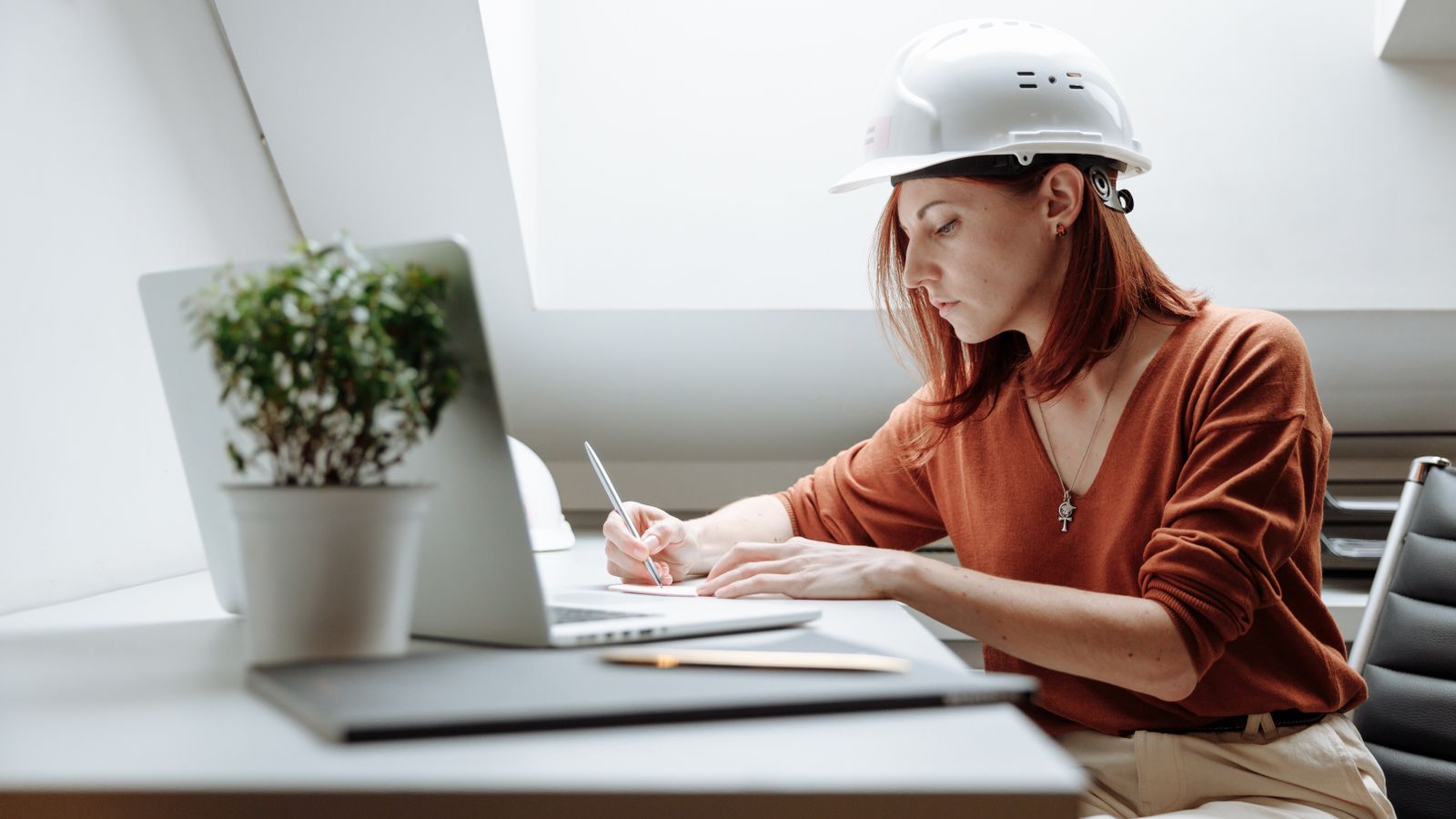 A person in a white hard hat writes notes at a desk beside a laptop.