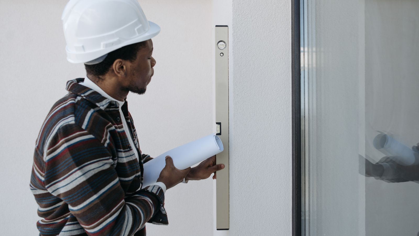 Un homme casqué vérifie un mur avec un niveau, un rouleau de plans à la main