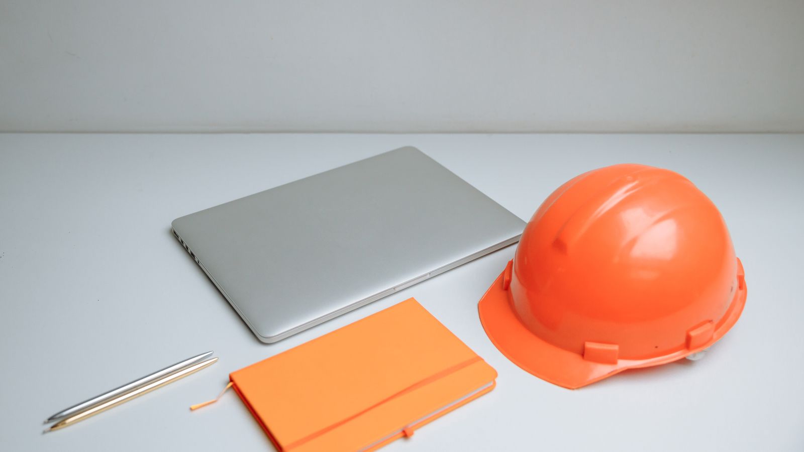 An orange hard hat, a closed laptop and an orange notebook on a white desk.