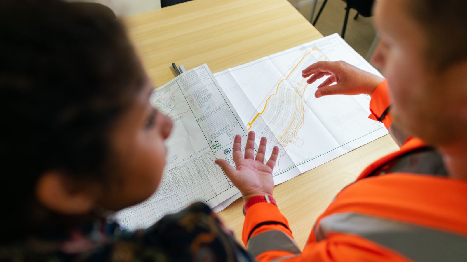 Two workers in high-visibility jackets review site plans spread across a wooden table.