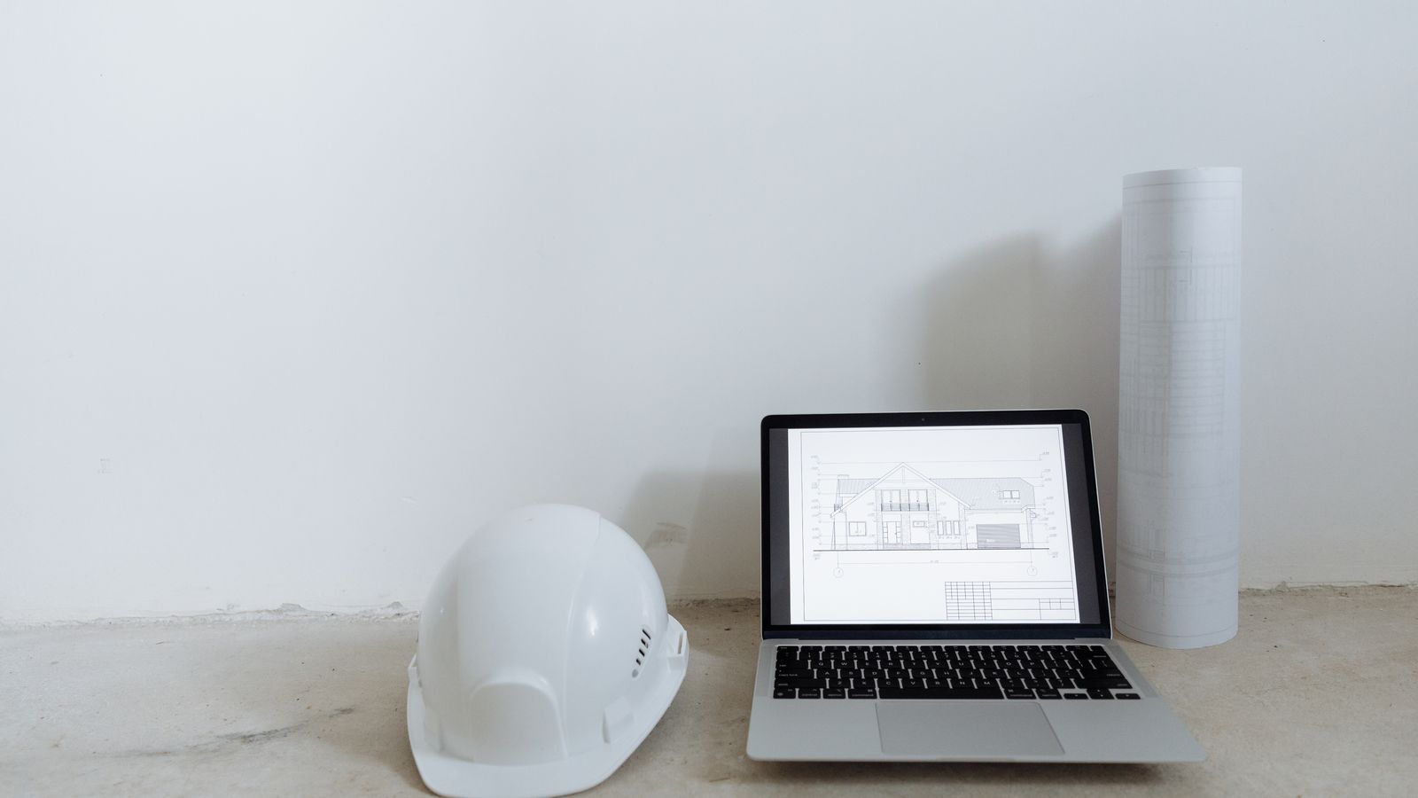 Casque blanc, ordinateur portable affichant des plans et rouleau de plans posés sur du béton.