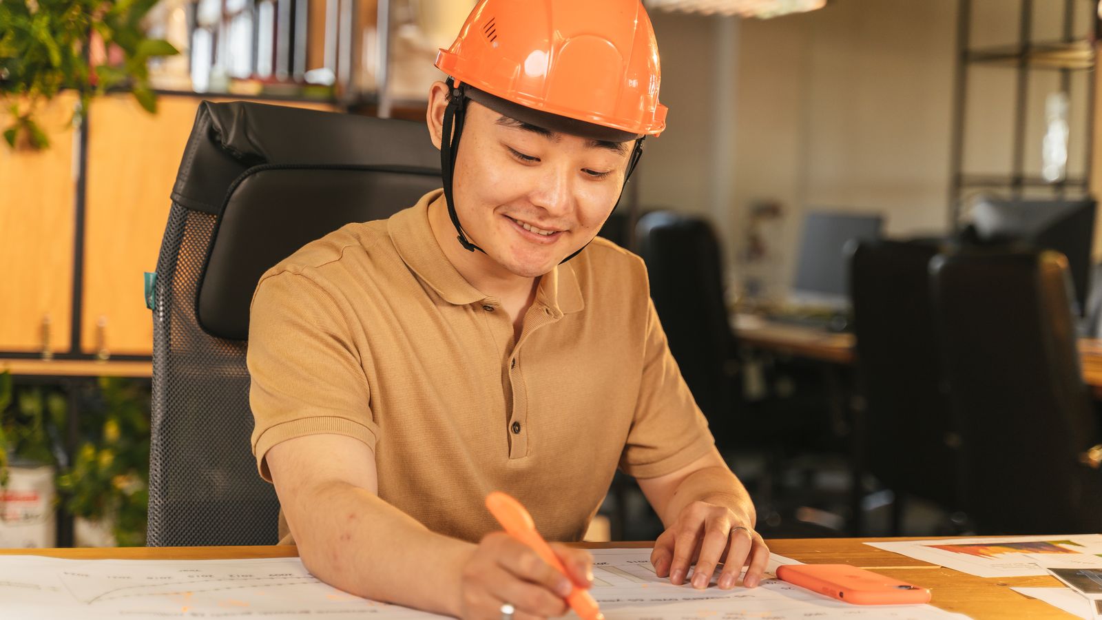 Un homme souriant coiffé d'un casque orange annote des plans à son bureau.