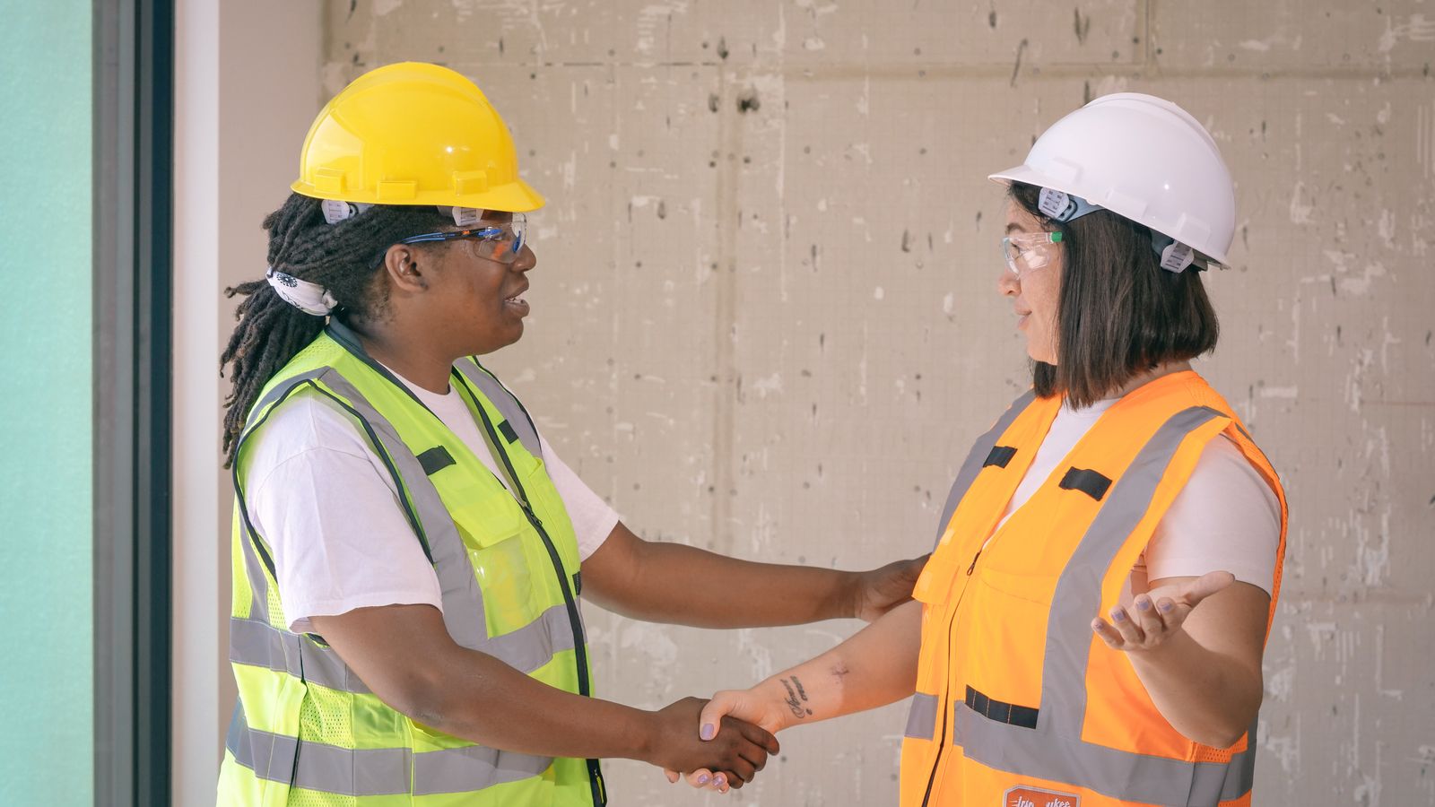 Two construction workers in hard hats, safety glasses and high-visibility vests shake hands indoors
