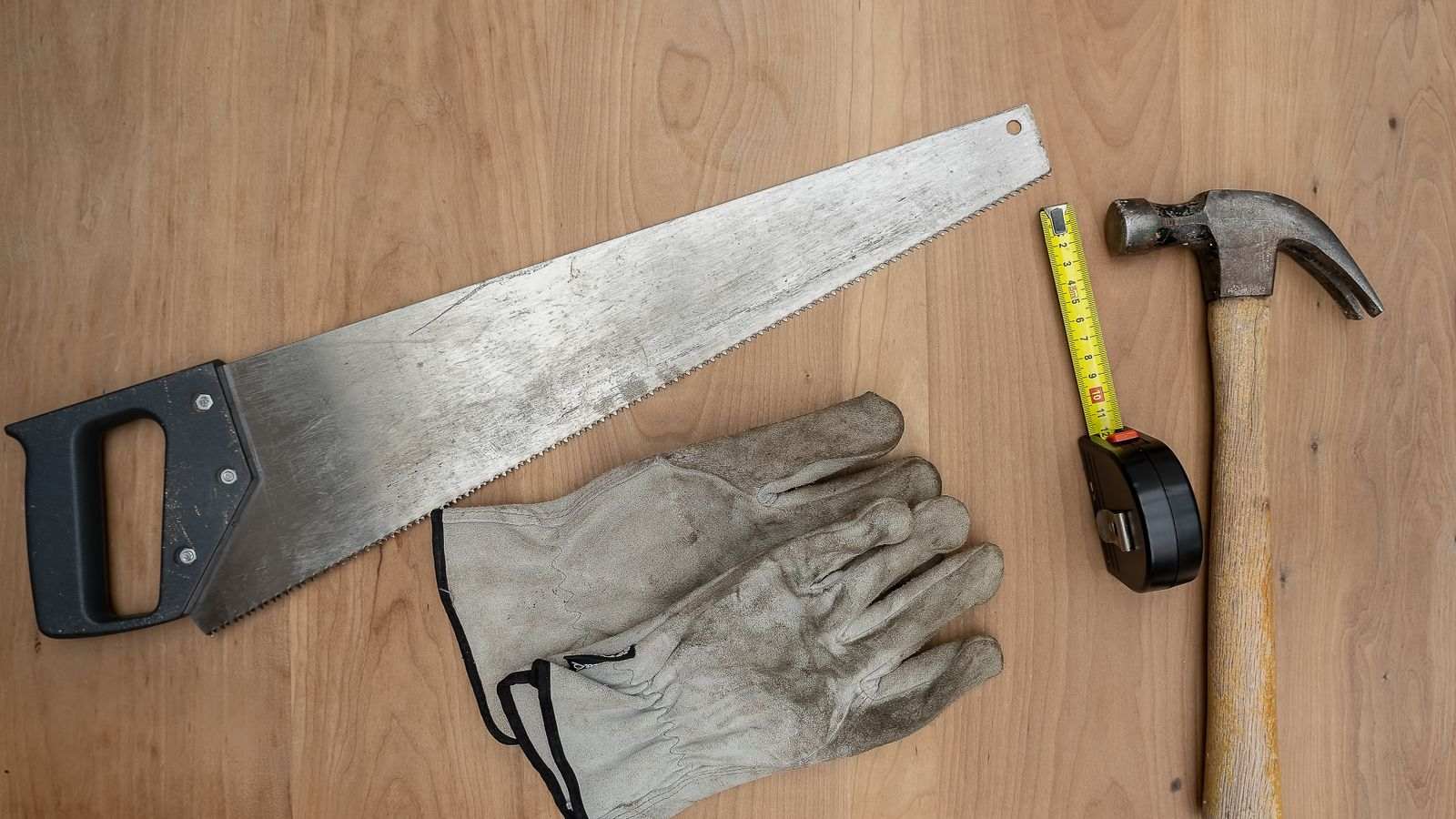 Scie, gants de travail, ruban à mesurer et marteau posés sur une table en bois.
