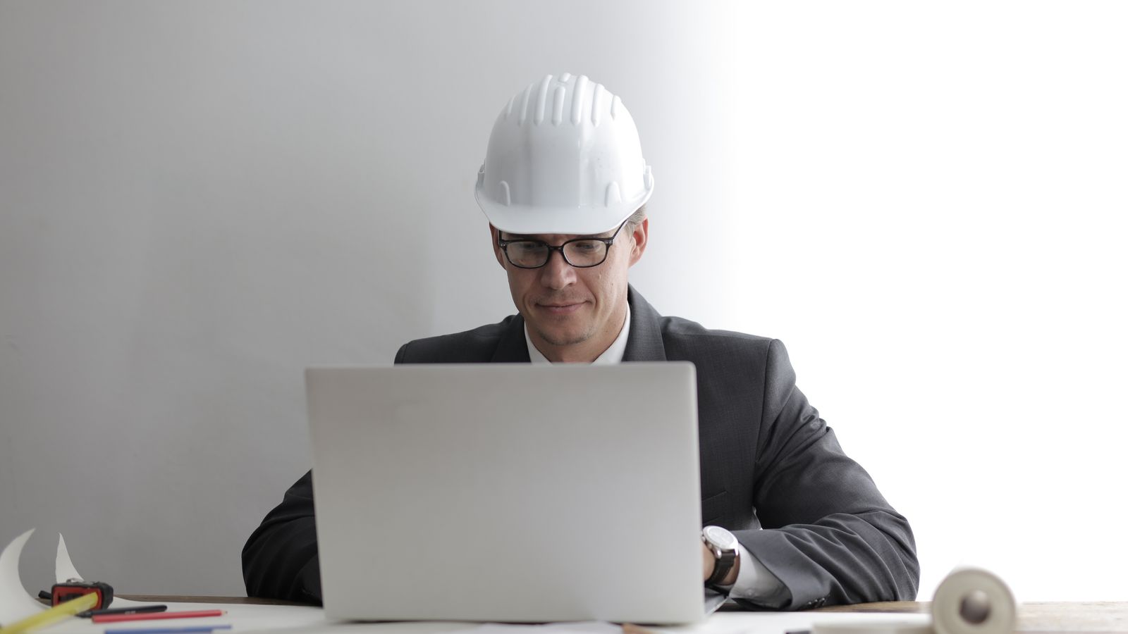 A contractor in a hard hat works at a laptop beside building plans in an office