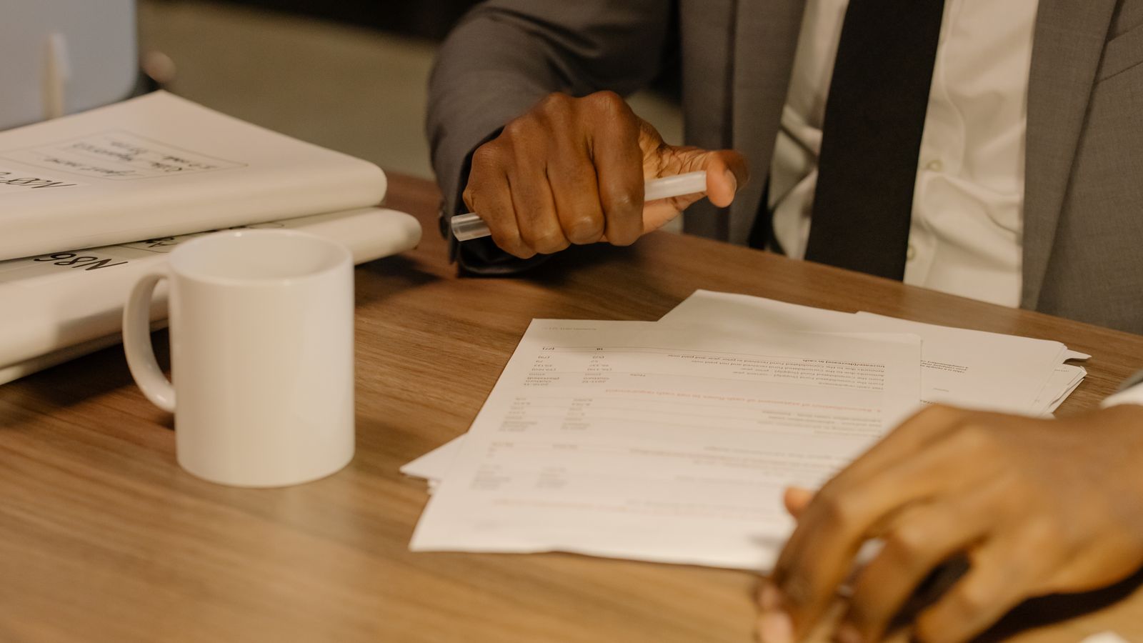 A person in a suit holds a pen over a stack of paperwork on a wooden desk.