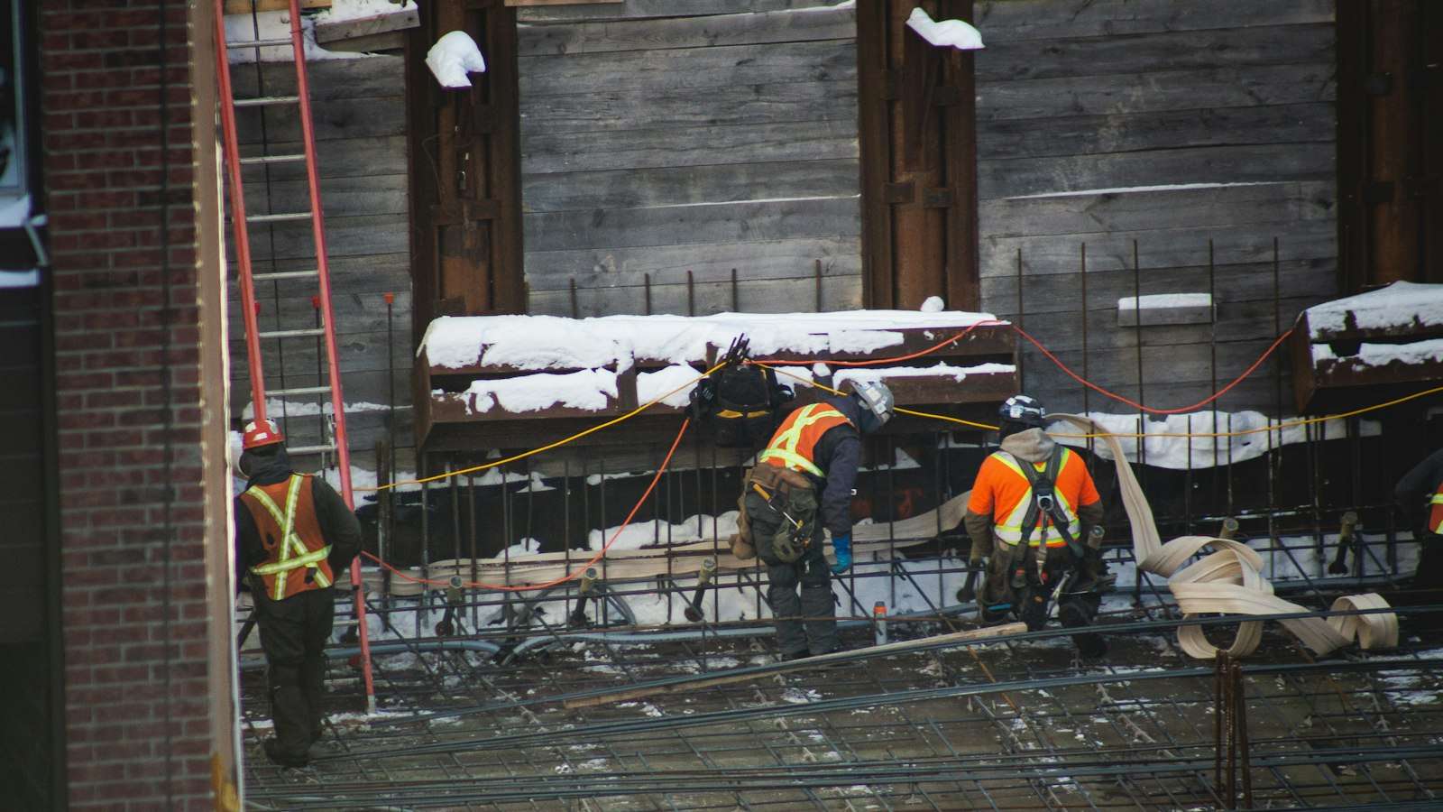 Workers in hard hats, vests and harnesses secured to lines on a snowy construction site